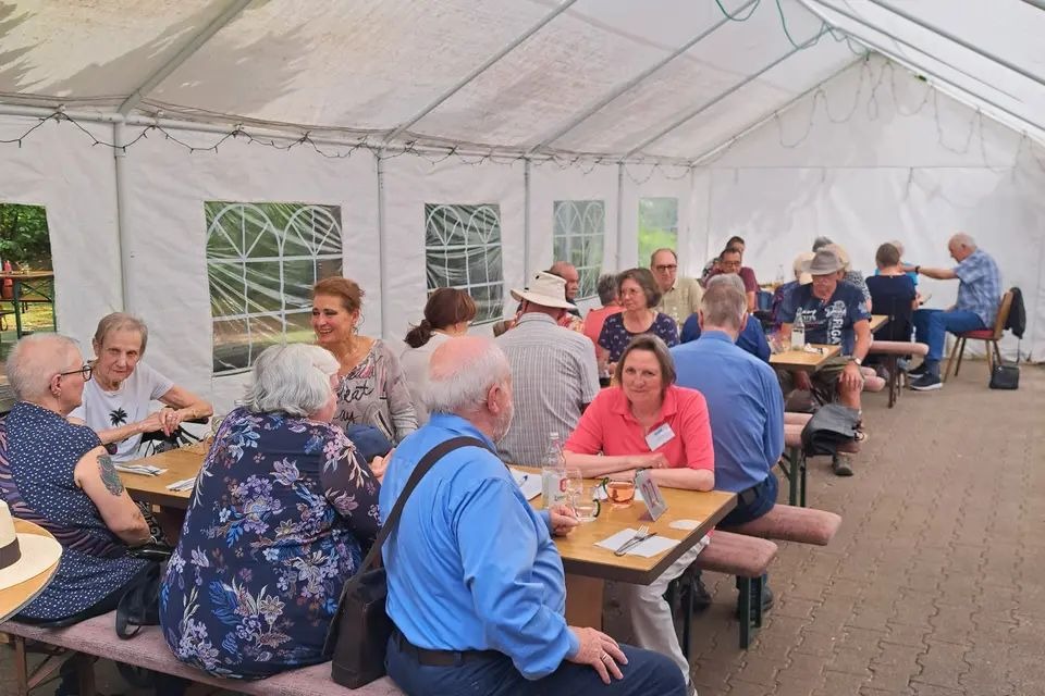 Es handelt sich um ein Foto von einer Gruppe &auml;lterer Menschen, die in einem gro&szlig;en wei&szlig;en Zelt sitzen und gemeinsam essen oder trinken. Das Zelt hat eine gew&ouml;lbte Decke aus leichtem, halbtransparentem Material und ist mit kleinen Lichterketten verziert. An den Seiten des Zelts sind gro&szlig;e, rechteckige Fenster angebracht &ndash; durch sie kann man auf B&auml;ume und Gr&uuml;nraum au&szlig;erhalb blicken. Die Besucher sitzen an langen Holztischen, die parallel zum Gang des Zelts aufgestellt sind. Jeder Tisch ist mit St&uuml;hlen oder B&auml;nken best&uuml;ckt, an denen sich mehrere Personen gegen&uuml;ber oder nebeneinander befinden. Die Kleidung der Anwesenden ist allt&auml;glich und bunt &ndash; manche tragen Hemden, andere Blusen oder Jacken. Einige haben Sonnenhut auf dem Kopf, was auf einen Sommertag hindeutet. Der Boden besteht aus grauen Pflastersteinen, die in einer regelm&auml;&szlig;igen Musterung verlegt sind. Die Atmosph&auml;re wirkt gem&uuml;tlich und gesellig.