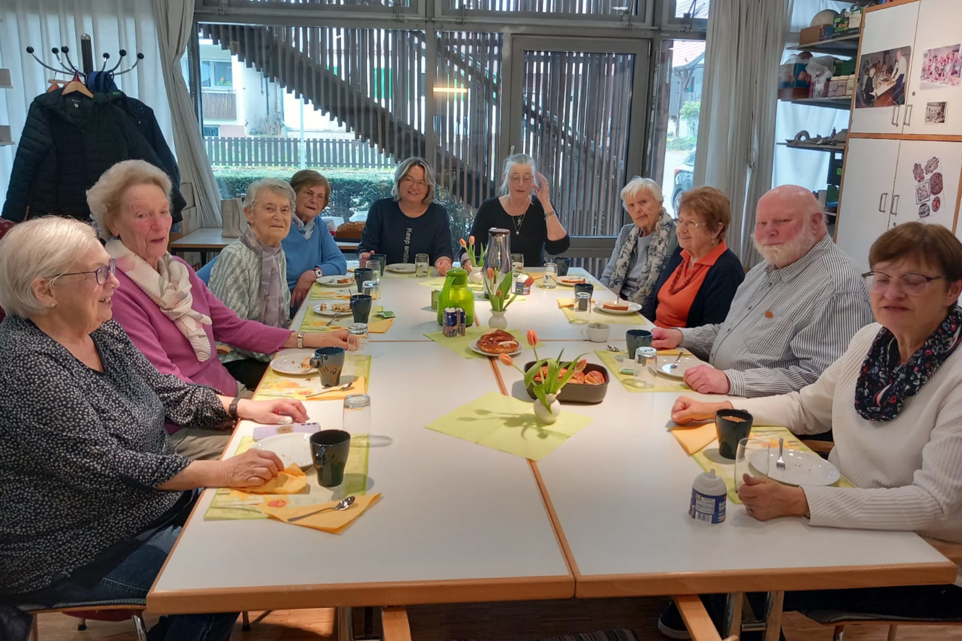 Neun Frauen und ein Mann bei einer geselligen Runde an einem gedeckten Tisch mit Kaffee und Kuchen.