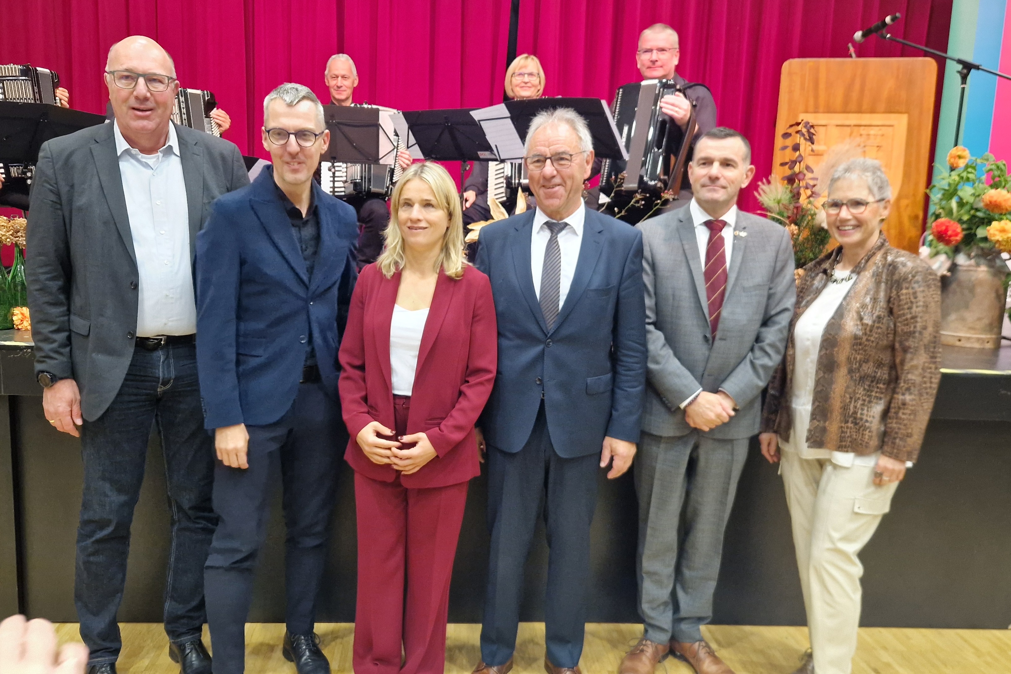 Gruppenbild: Ehreng&auml;ste stehen vor der B&uuml;hne. im Hintergund dunkelroter Vorhang, davor auf der B&uuml;hne das _Akkordeonensembel
