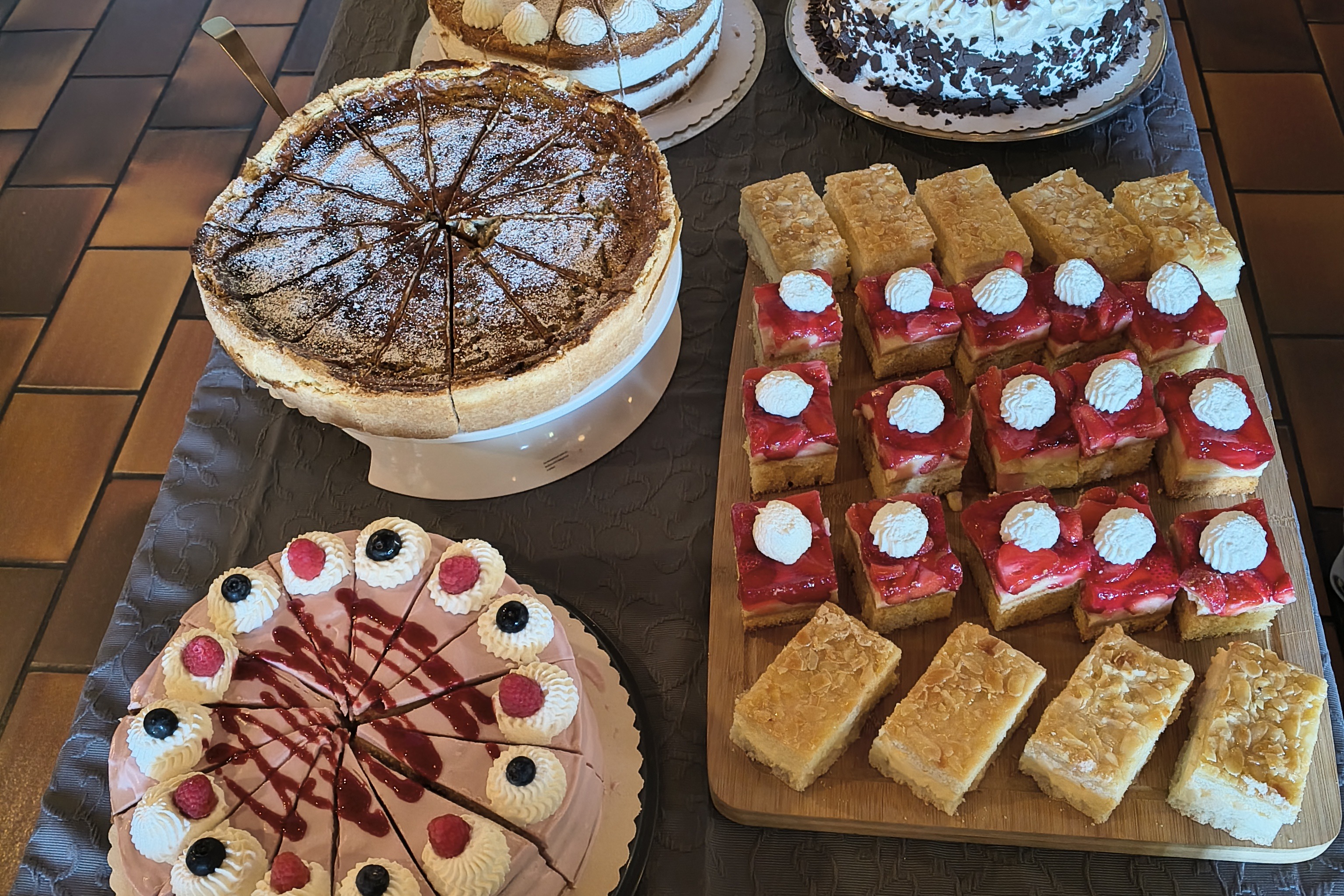 Mehrere verschiedene Kuchen und Torten auf einem dunklen Tisch, darunter runde Torten mit Sahne und Beeren sowie rechteckige Kuchenst&uuml;cke mit roten und gelben Bel&auml;gen.