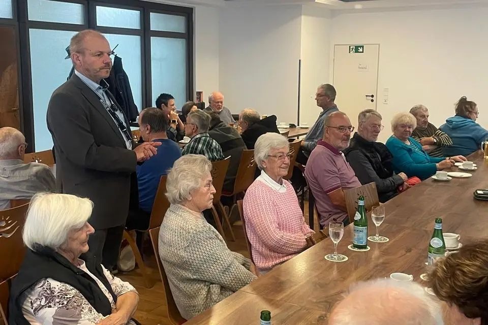 Ein &auml;lterer Mann steht im Vordergrund einer Versammlung in einem hellen Raum und h&auml;lt eine Pr&auml;sentation. Mehrere &auml;ltere Personen sitzen an einem langen Holztisch, h&ouml;ren zu oder unterhalten sich leise. Auf dem Tisch stehen Gl&auml;ser, Tassen und Flaschen mit Getr&auml;nken. Im Hintergrund sind weitere G&auml;ste an anderen Tischen zu sehen, einige stehen oder bewegen sich. Durch ein gro&szlig;es Fenster links dringt diffuses Tageslicht herein, w&auml;hrend die W&auml;nde wei&szlig; sind und eine T&uuml;r mit einem gr&uuml;nen Notausgangsschild im Hintergrund sichtbar ist. Die Atmosph&auml;re wirkt entspannt und gemeinschaftlich, wie bei einem Treffen oder einer Veranstaltung in einem Gemeinschaftszentrum.