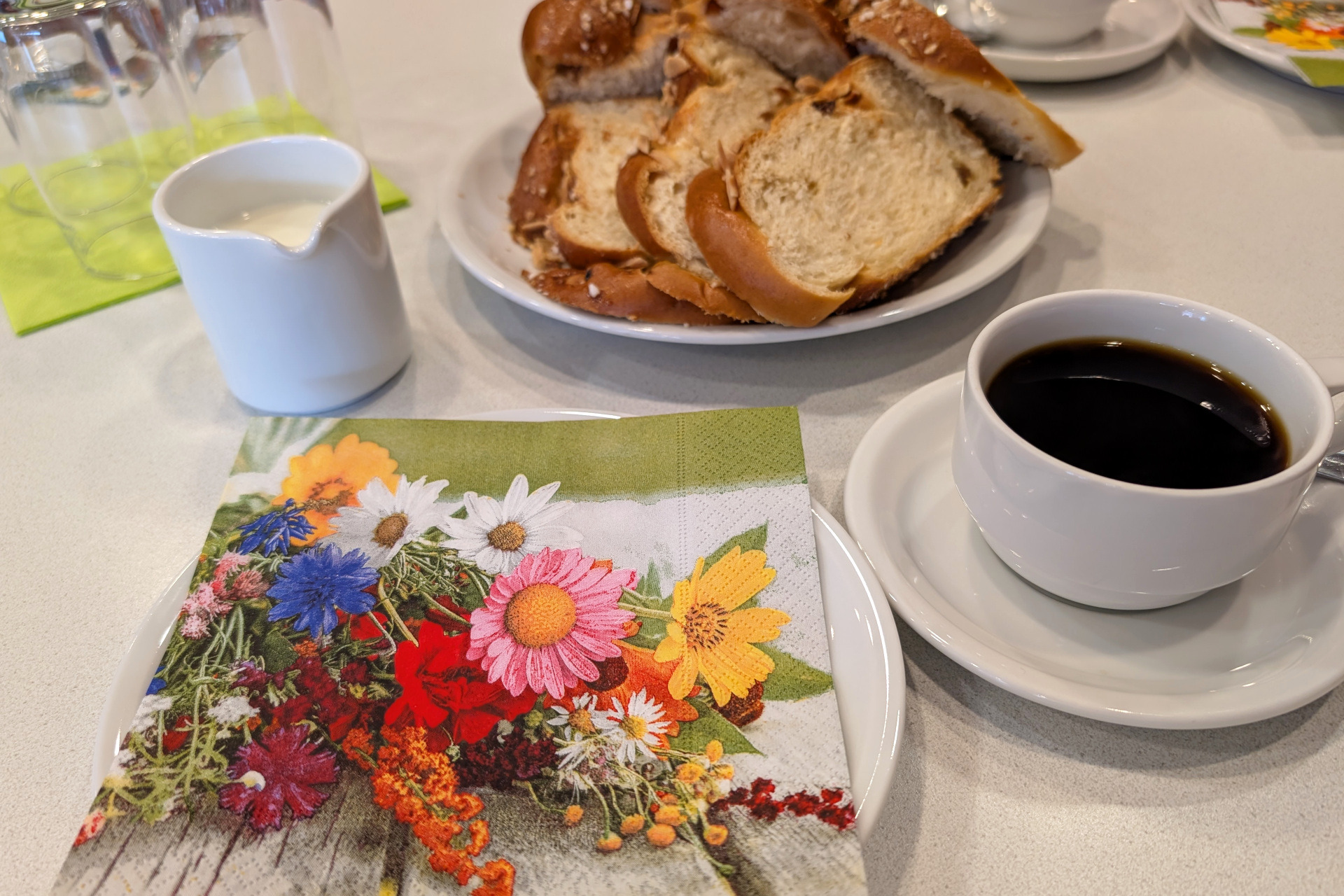 Kaffee-Nachmittag Blumenserviette auf einem Teller, daneben eine Tasse Kaffee, ein Milchkännchen und ein Teller mit frischem Hefezopf.