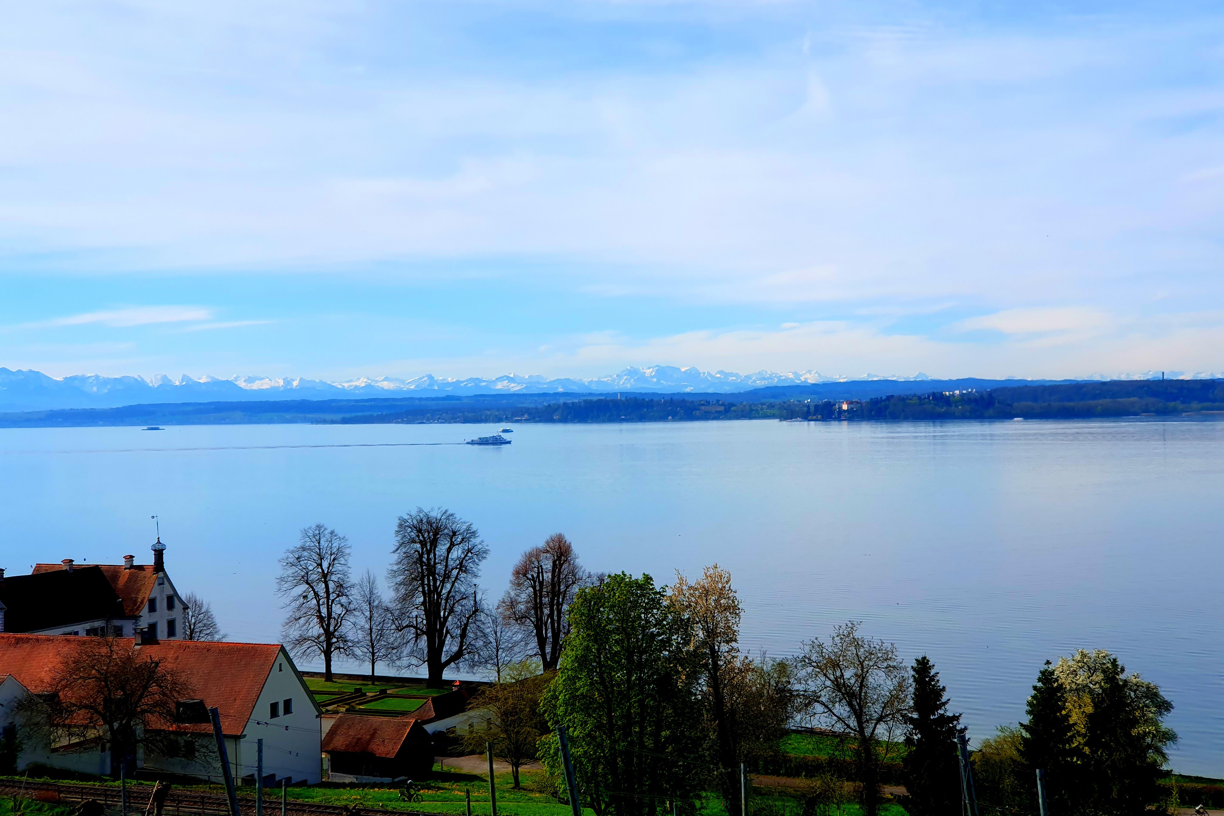 Tagesausflug 2 Ein grosser See, im Vordergrund das Ufer mit Bäumen und zwei Häuser, am gegenüber liegenden Ufer erkennt man die Alpen.