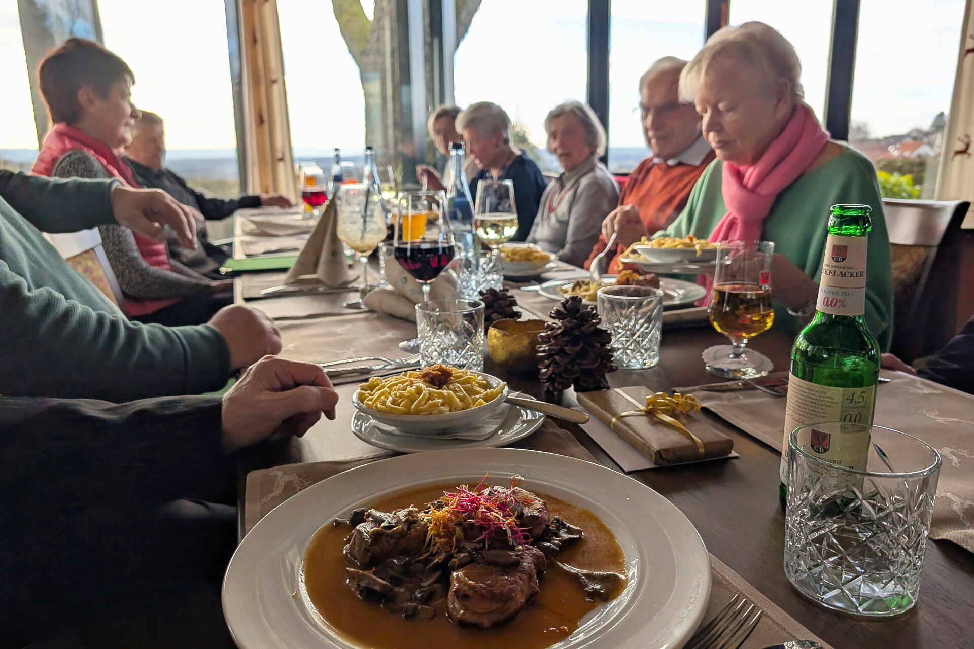 Tisch, an dem mehrere Personen sitzen. Im Vordergrund ein Teller mit Fleisch und Soße, daneben ein kleiner Teller mit Spätzle. Im Hintergrund etwa zehn Personen, leicht verschwommen, die sich unterhalten.