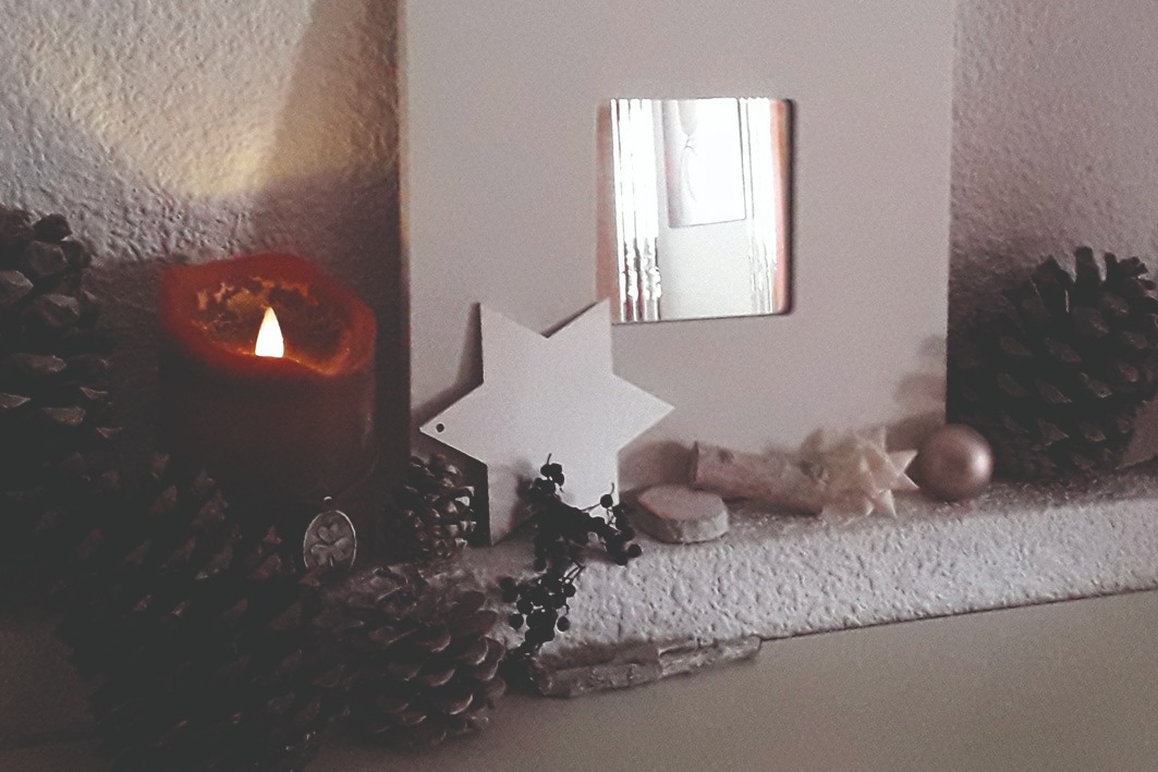 Links brennende Kerze mit Lichtschein nach oben. Daneben Weihnachtsdeko