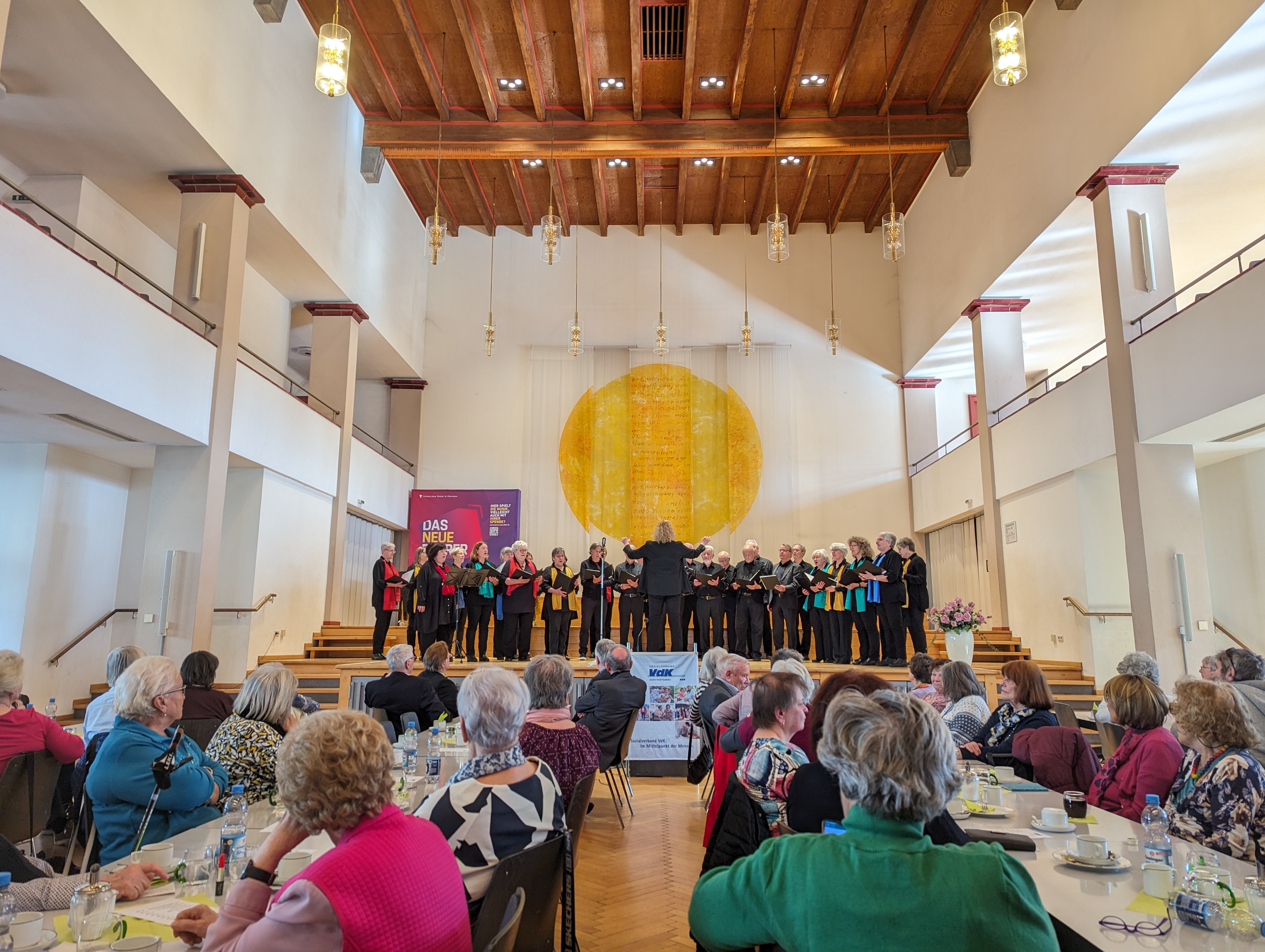 Panorama vom Festsaal. Mit mehreren Tischreihen, an den viele Leute sitzen. Im Hintergrund die Bühne mit Chor.