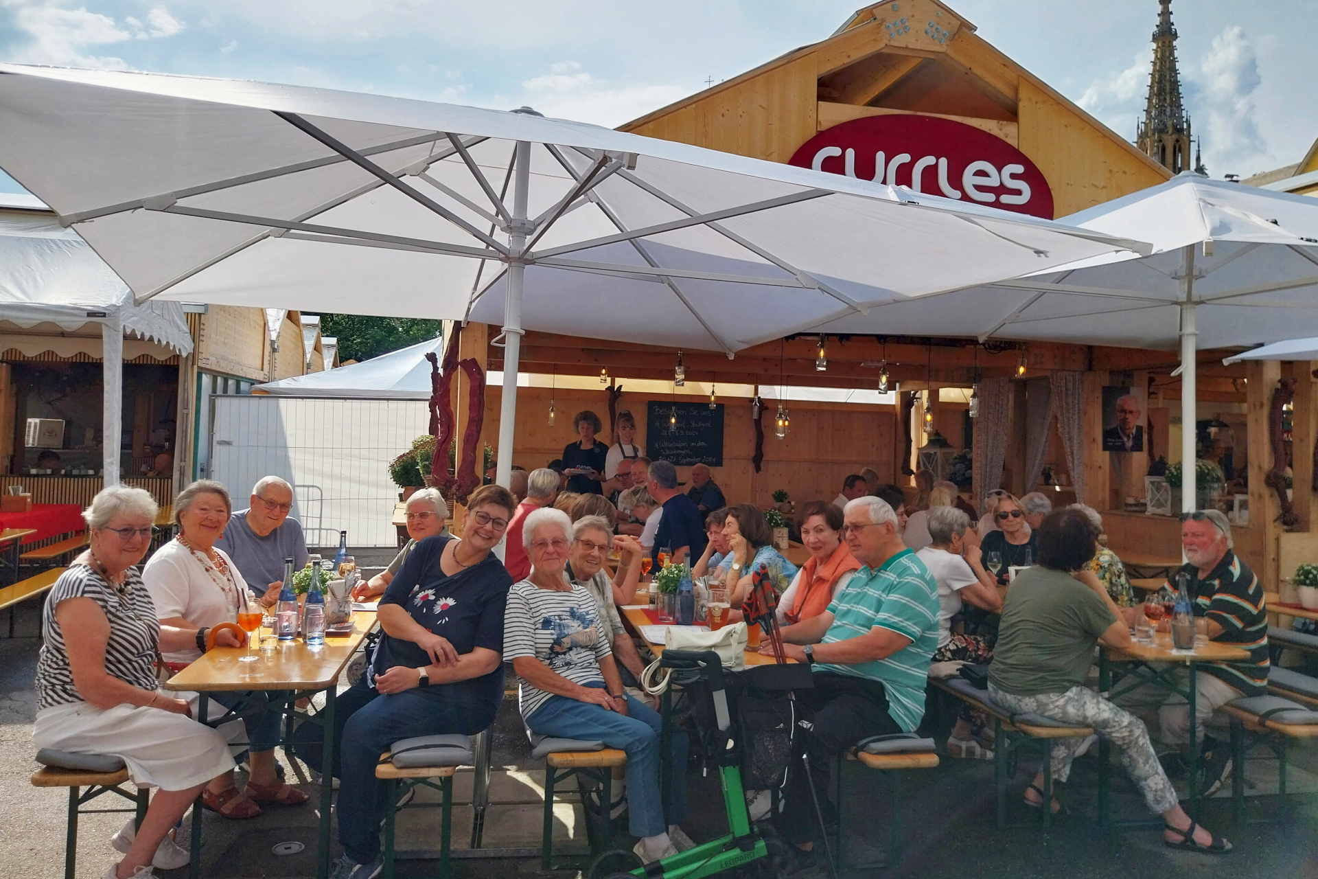 ESTiVAL. Eine Gruppe von Rentnern sitzt an Bierzeltgarnituren und lächelt in die Kamera. Im Hintergrund steht eine Holzhütte mit einem Schild, auf dem "Currles" zu lesen ist.