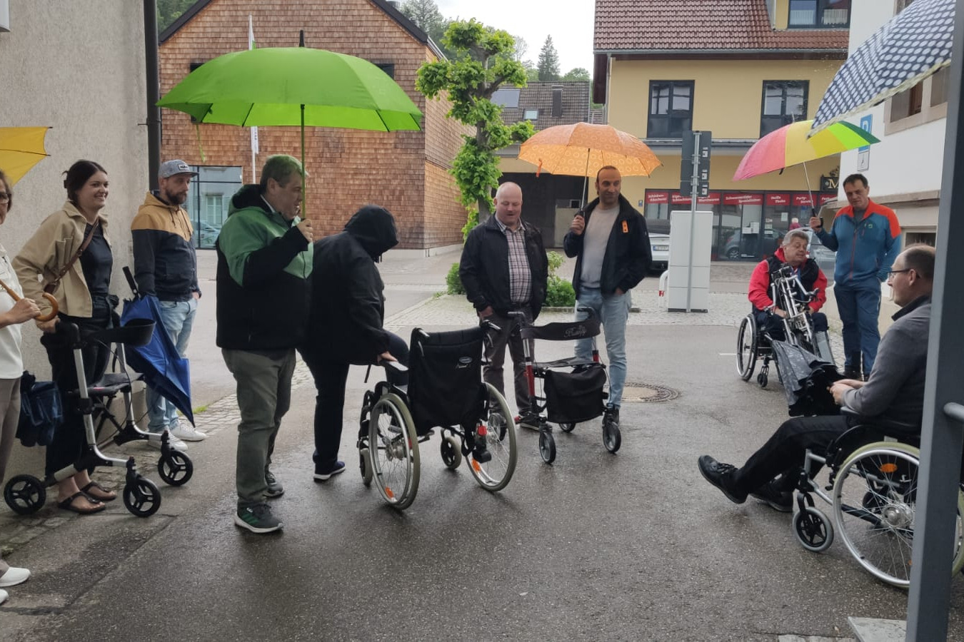 VdK-Städtlirunde mit Rollstühlen, Schirmen usw. Für viele eine neue Erfahrung, ein noch zu besetzender Rollstuhl, einige schon besetzte Rollstühle, eine Person am Rollator, mehrere Menschen mit Schirmen vor dem neuen MTB-Haus