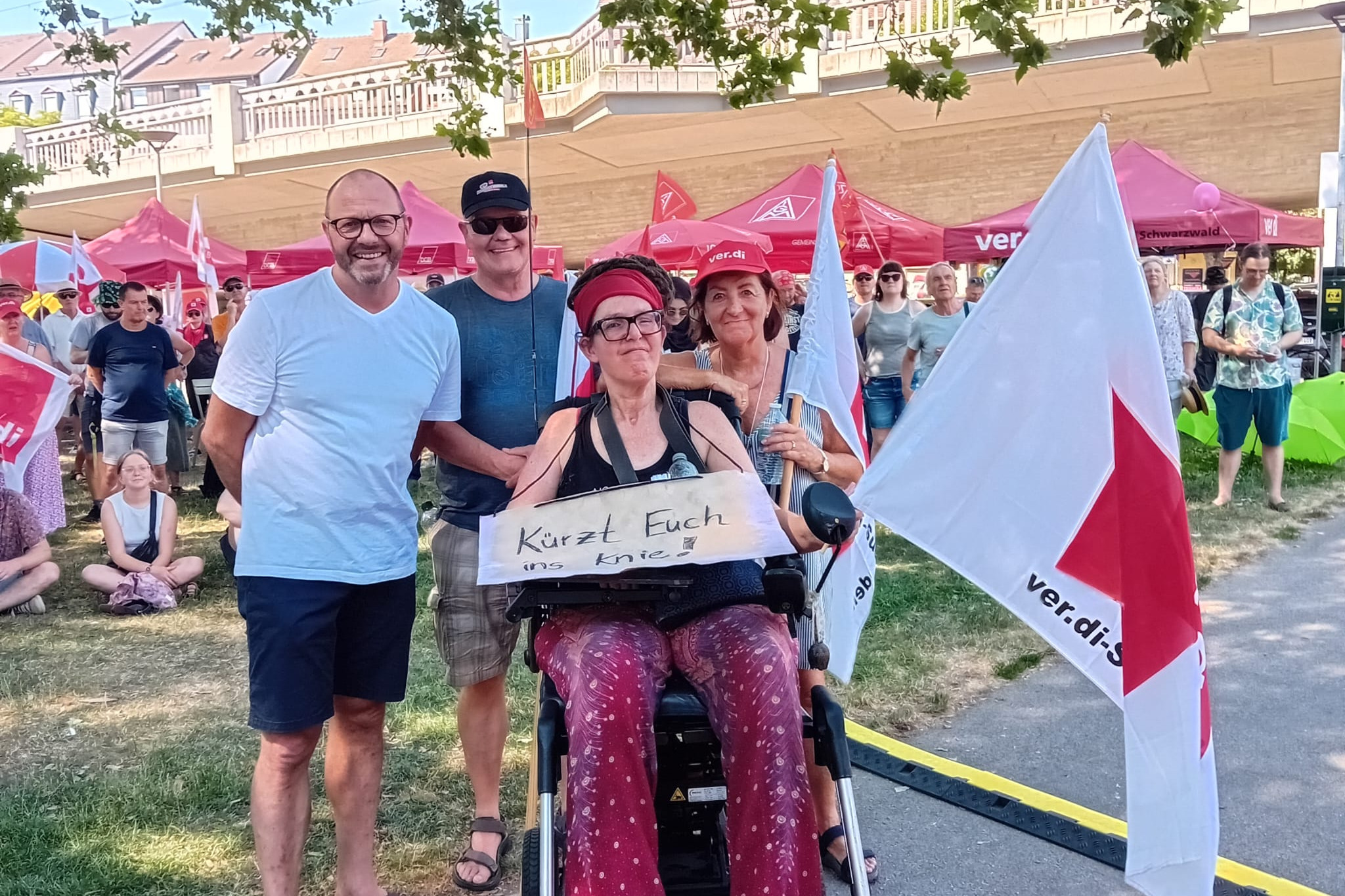 Freiburgs Sozialb&uuml;rgermeister Roland Meder, Schatzmeister Andreas Miller, VdK Dreisamtal, stehend von links Homepagebeauftragte Karin Maier im Elektrorollstuhl, mit umh&auml;ngendem Pappschild "K&uuml;rzt euch ins Knie" und ver:di-Fahne, Ortsvorsitzende Brigitte Mauz, stehend, bei der Kundgebung im St&uuml;hlinger-Park in Freiburg am 27.6.2026