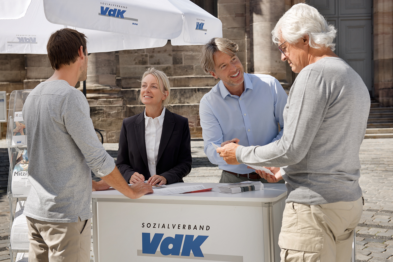 Eine freundliche Frau und ein freundlicher Mann stehen hinter einer VdK-Werbetheke und einem VdK-Schirm auf einem Marktplatz, zwei Männer lassen sich von den beiden beraten