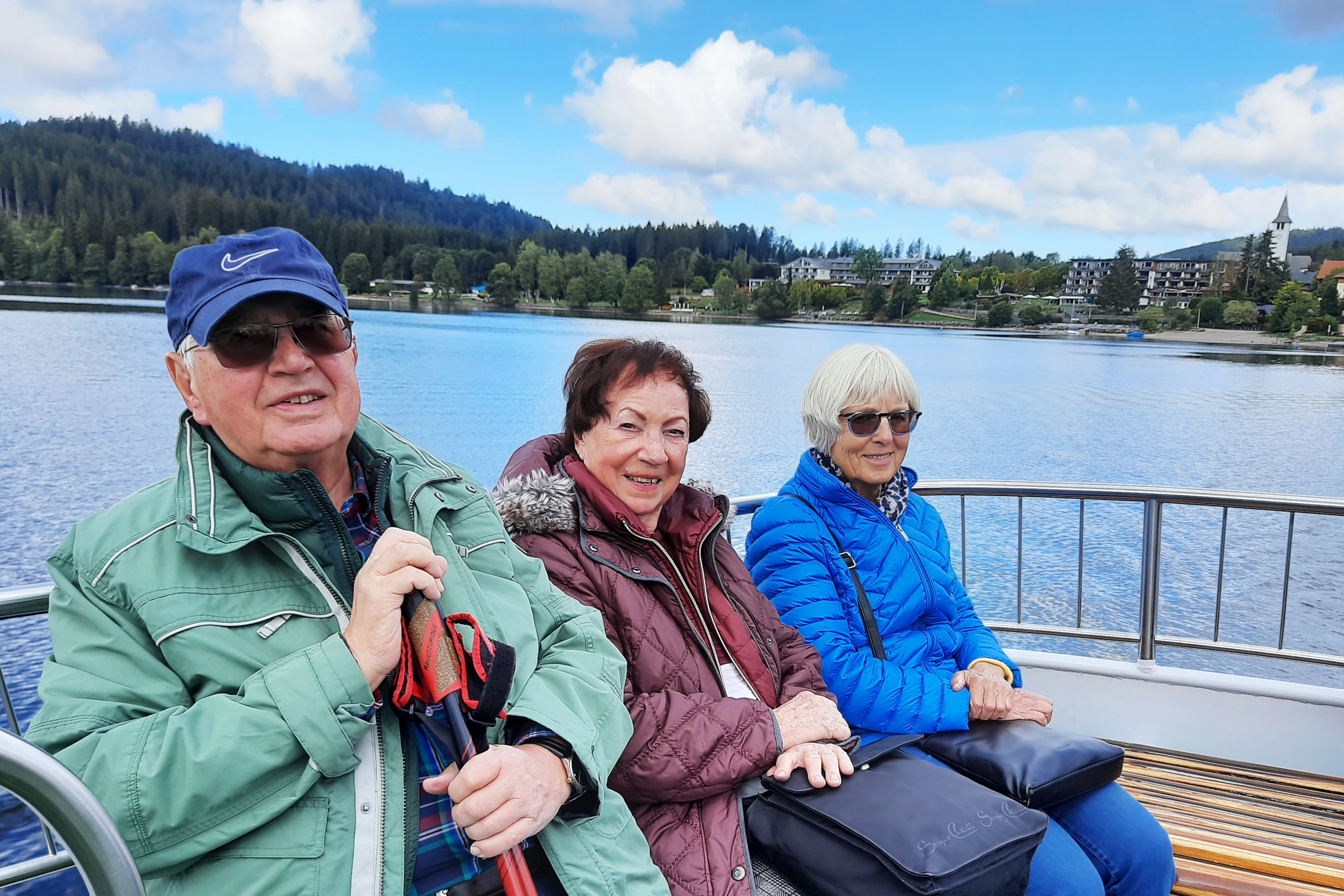 Titisee Drei ältere Menschen sitzen auf dem Oberdeck eines Schiffs. Im Hintergrund sind der Titisee, Wald und einige Gebäude am Ufer zu sehen.