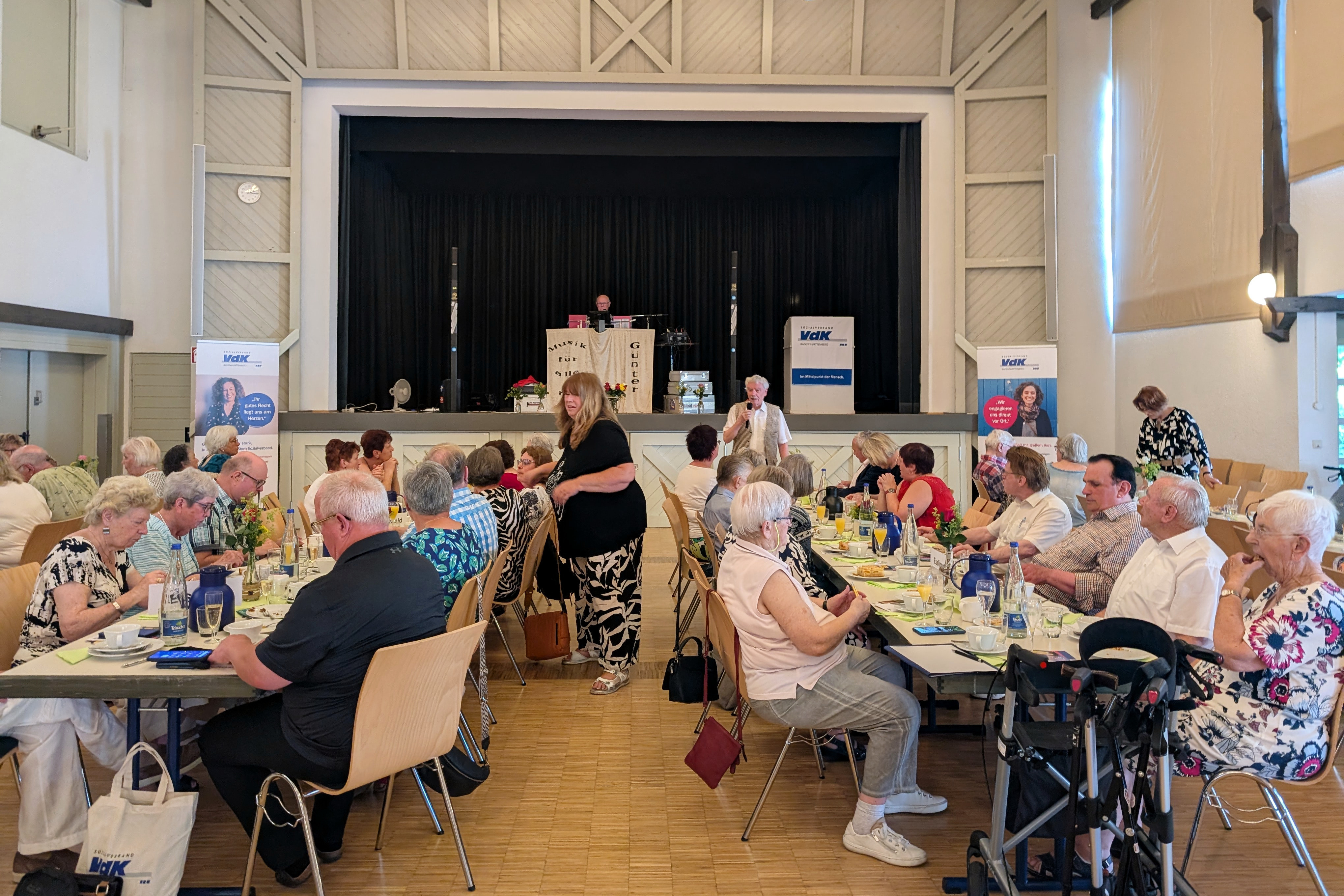 Sommerfest Großer Festsaal mit mehreren Tischreihen, an denen vorwiegend ältere Menschen sitzen. Im Hintergrund befindet sich eine Bühne, auf der ein DJ an seinem aufgebauten Equipment sitzt.
