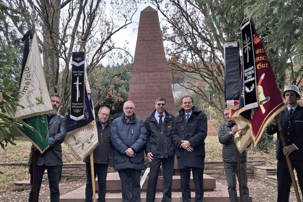 Vor dem Krigs Denkmal stehen vier Fahne Tr&auml;ger von Sch&uuml;tzenverein, MGV, S&auml;ngerbund, Tsg, Wilhelmsfeldfreiwillige Feuerwehr Wilhelmsfeld und der zweite Vorsitzende des VDK Klaus Rehberger, der erste Vorsitzenden Michael Reinhard und B&uuml;rgermeister Dr. Tobias Dangel