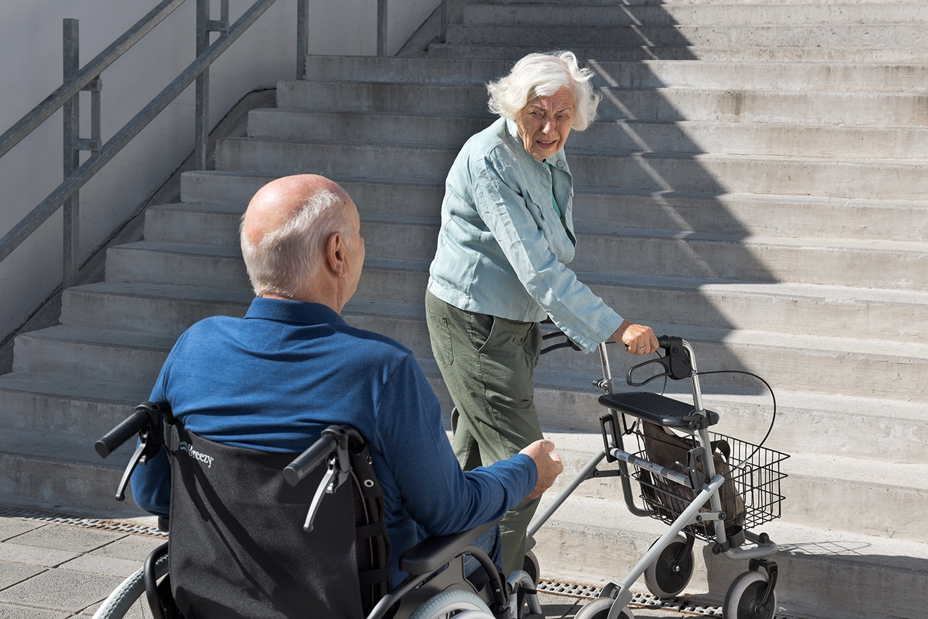 Ein Rollstuhlfahrer und eine Dame mit Rollator vor einer Treppe Ein Rollstuhlfahrer und eine Dame mit Rollator vor einer Treppe