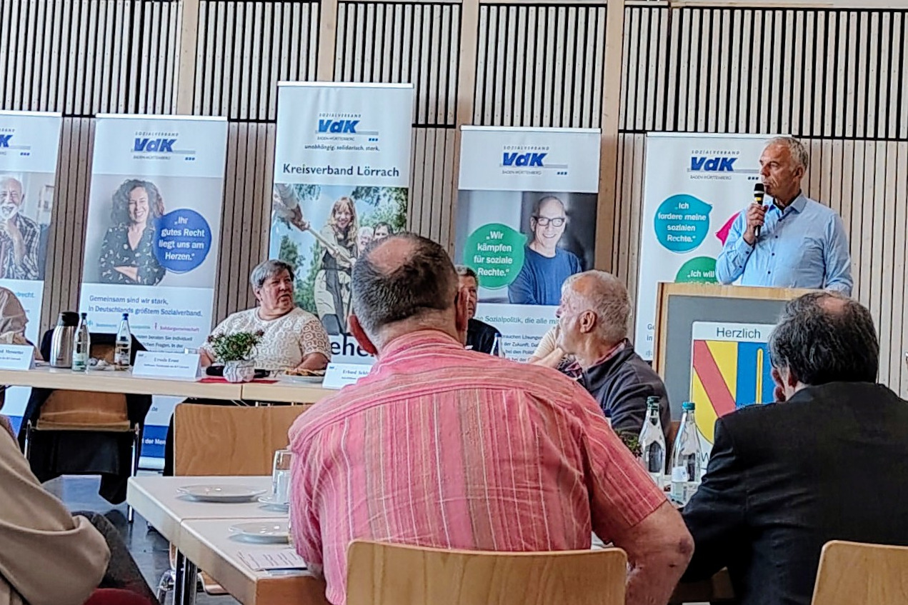 Der Kreisvorsitzende Manfred Merstetter bei der Kreisdelegiertenkonferenz am Rednerpult, daneben das Podium mit den Ehrenamtlichen des Kreisverbandes, dahinter mehrere Roll-ups des VdK, die Zuh&ouml;renden an Tischen, einige Zuh&ouml;rende