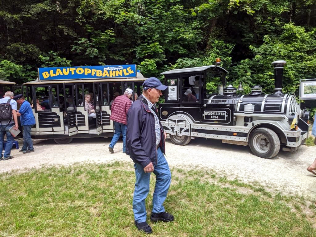 Mehrere Rentner stehen vor einer Touristenbahn, die wie eine kleine Dampflok aussieht; in den offenen Waggons des Zuges sitzen bereits mehrere Personen.