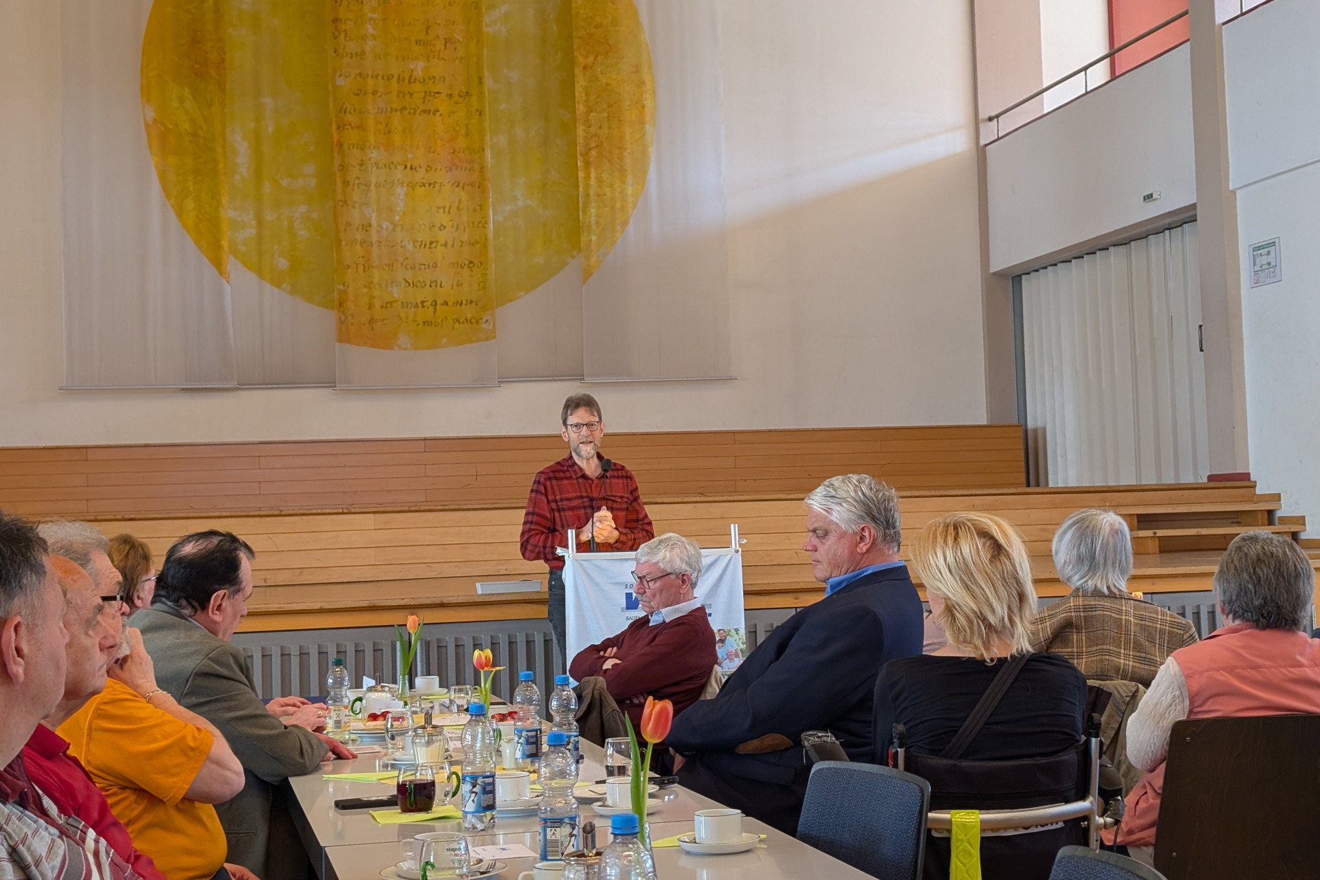 Im Vordergrund eine Tischreihe mit sitzenden Menschen, die aufmerksam dem Redner am Pult vorne lauschen.