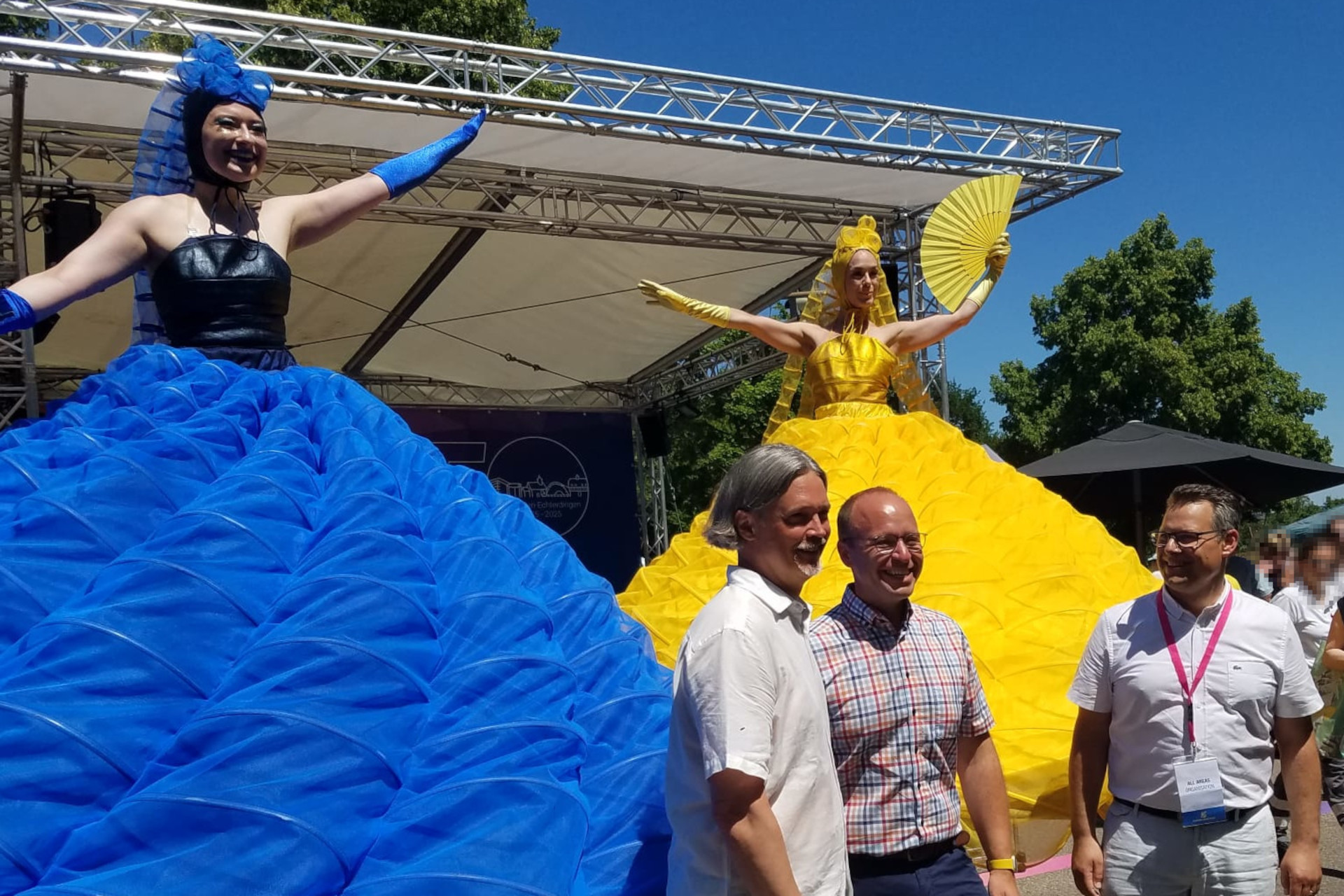 Stadtfest Leinfelden-Echterdingen Zwei Frauen auf einer Bühne, eine in einem strahlend blauen Kleid mit weitem, aufgebauschtem Rock, die andere in einem gelben, ebenfalls aufgebauschten Rock. Vor der Bühne stehen drei Männer.