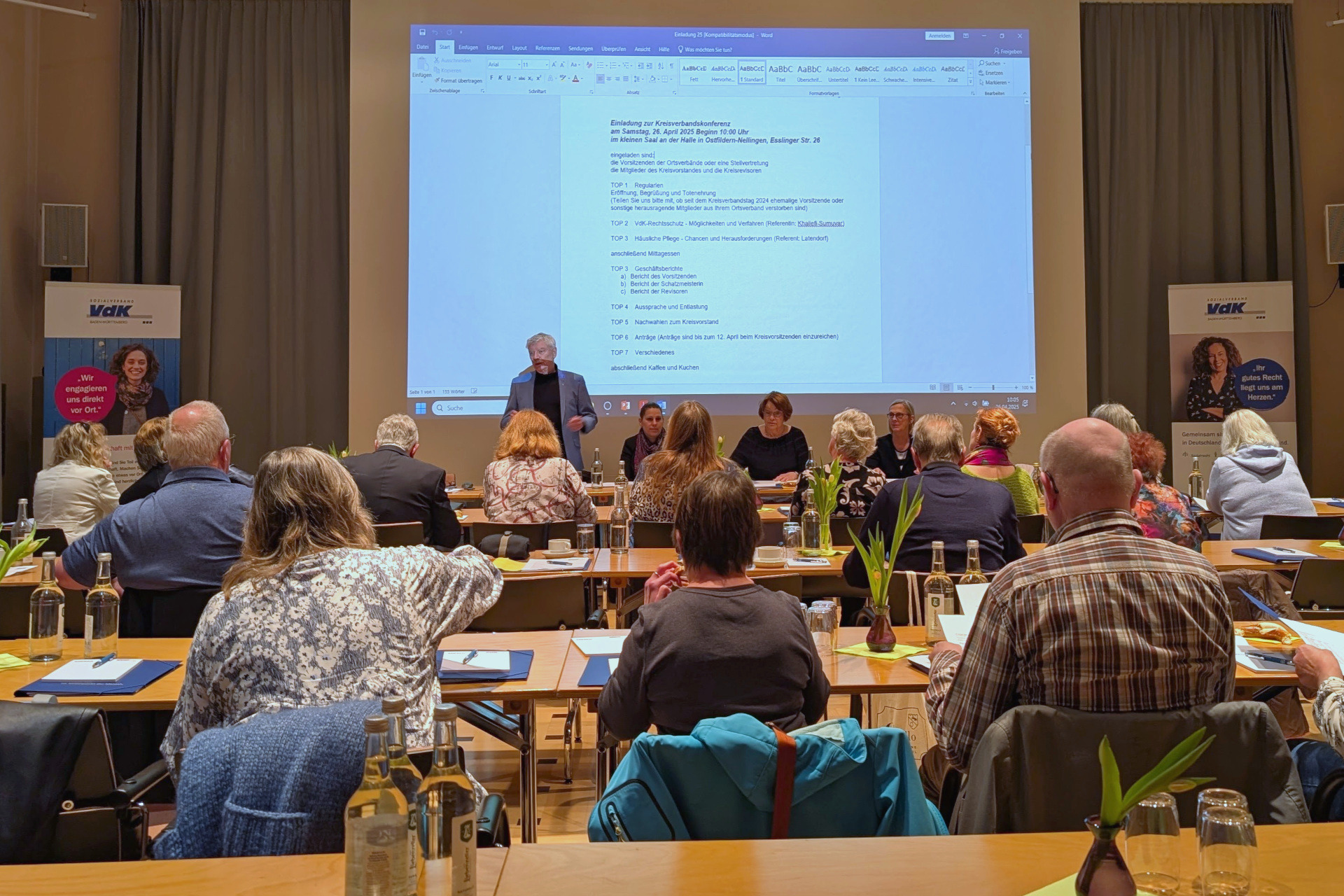Kreisverbandskonferenz Blick in den Vortragssaal von hinten auf mehrere Tischreihen mit Konferenzteilnehmenden; vorne zeigt eine Leinwand die Tagesordnungspunkte, davor sitzt der geschäftsführende Vorstand des Kreisverbands.