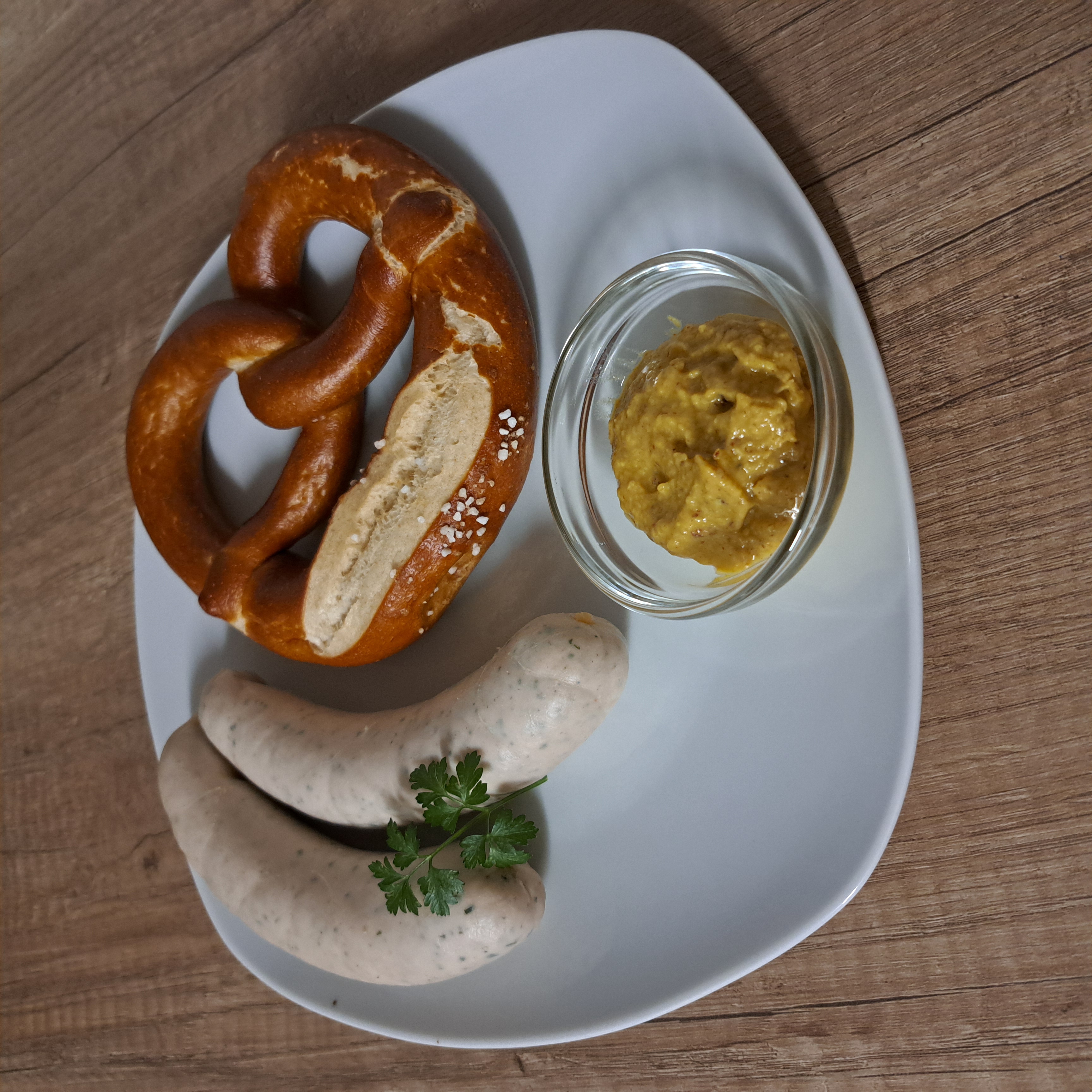 Zwei Weißwürste mit Petersilie, eine Brezel und ein Schälchen mit süßem Senf auf einem weißen, eckigen Teller