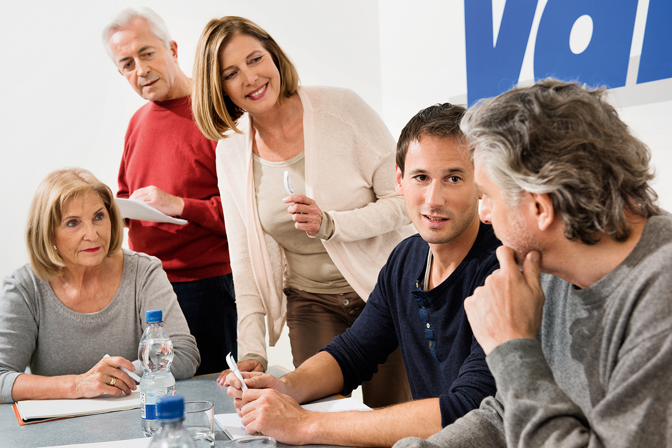 Drei Menschen sitzend am Tisch, zwei stehend, miteinander sprechend