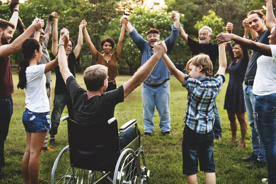 Gruppe fröhlicher Menschen verschiedenen Alters, verschiedener Ethnie, mit und ohne Handicap stehen im Park im Kreis. Halten sich an den Händen, strecken Hände in die Luft.