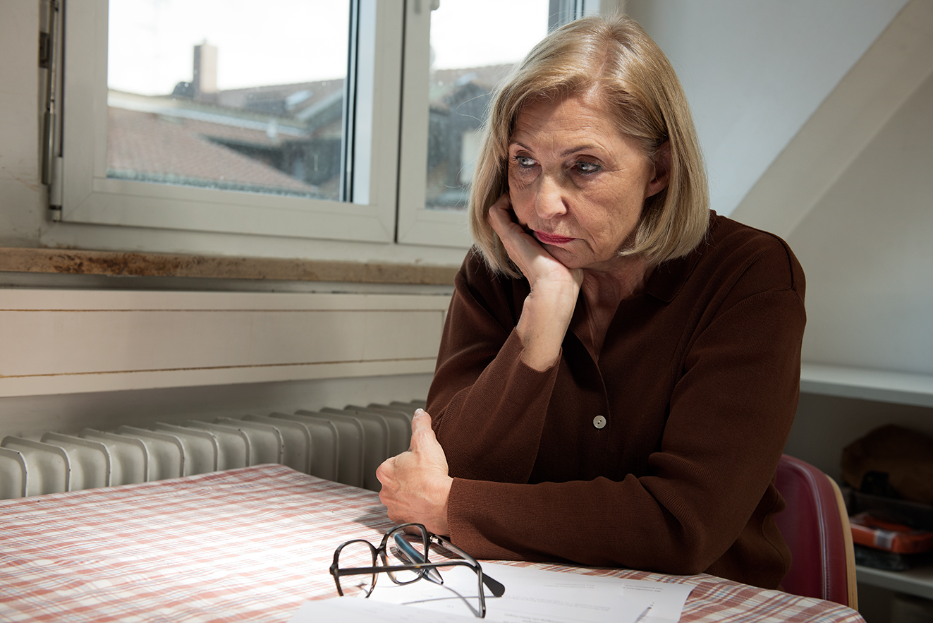 Eine verzweifelt aussehende Frau sitzt an einem Tisch, ihre Brille liegt auf dem Tisch und daneben einige Papiere