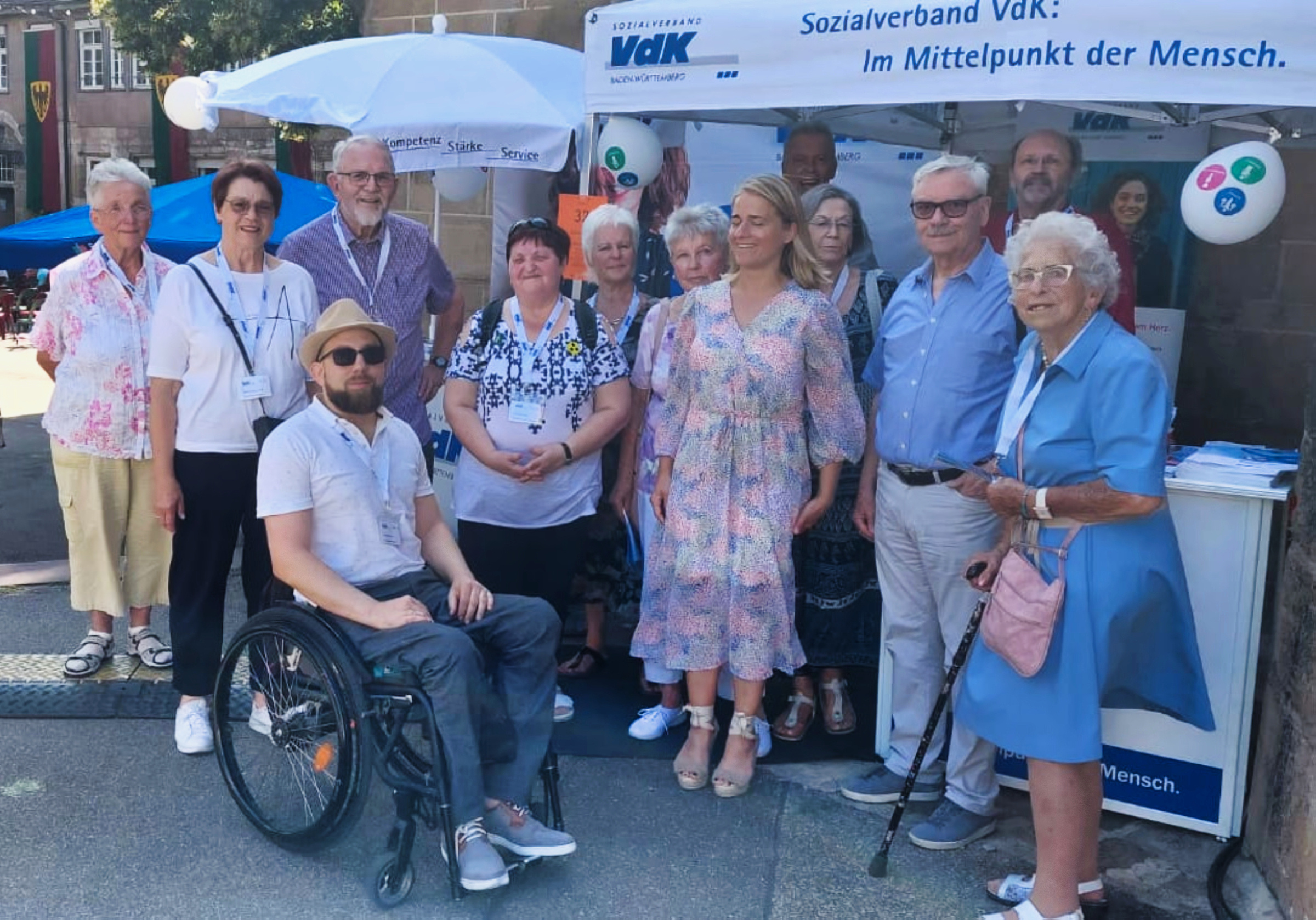 Gruppenbilder von 13 Personen vor einem Infostand mit VdK-Werbung. Unter der Gruppe Verena Bentele in einem Sommerkleid.