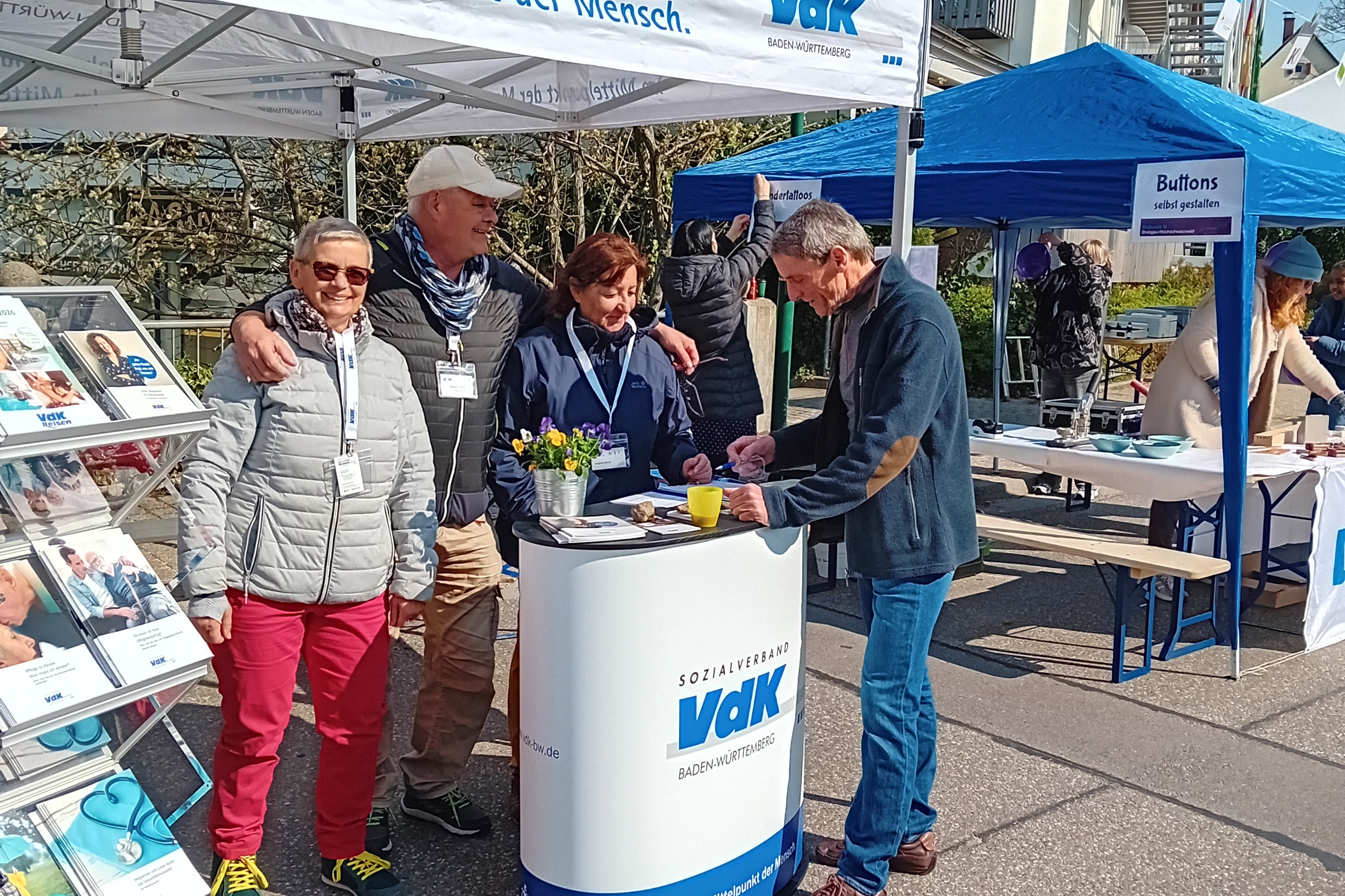 Die Schriftf&uuml;hrerin Gabi Morath, der Schatzmeister Andreas Miller und die Ortsvorsitzende Brigitte Mauz stehend hinter dem VdK-Infostand mit einem die strahlende Fr&uuml;hlingssonne nutzenden und den "Infospeicher" auff&uuml;llenden Besucher. Links steht ein Brosch&uuml;renst&auml;nder.